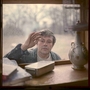 Man with a curious expression touches a dusty window from outside, viewed from inside a room with books and a decorative lamp.