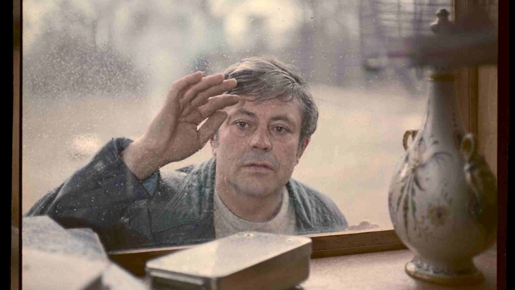 Man with a curious expression touches a dusty window from outside, viewed from inside a room with books and a decorative lamp.