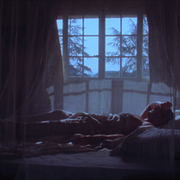 In a dimly lit bedroom, a person lies on a bed beneath a translucent mosquito net canopy. Blue moonlight enters through a large-paned window behind them, casting soft shadows across the room. Silhouettes of trees are visible outside, while the figure rests with one arm bent behind their head, creating a quiet, contemplative atmosphere. The image has a dreamlike, cinematic quality, with deep blues and soft focus enhancing the sense of solitude and reflection. 