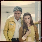 Man in yellow jacket and woman in brown and yellow top standing close together, looking thoughtfully off-camera.