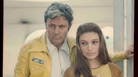 Man in yellow jacket and woman in brown and yellow top standing close together, looking thoughtfully off-camera.