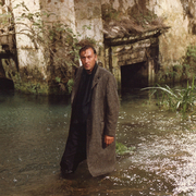 Man in a long coat standing in shallow water near an old stone structure with overgrown plants.