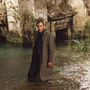 Man in a long coat standing in shallow water near an old stone structure with overgrown plants.