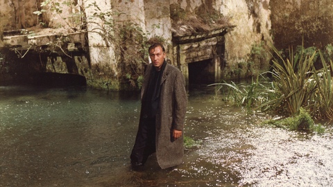 Man in a long coat standing in shallow water near an old stone structure with overgrown plants.