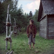 A barefoot boy in rustic clothes walks outside a wooden cabin, surrounded by green grass and birch tree fencing.