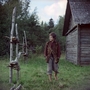A barefoot boy in rustic clothes walks outside a wooden cabin, surrounded by green grass and birch tree fencing.