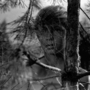 Black and white photo of a boy’s face partially obscured by pine branches and a large spider web in a forest setting.