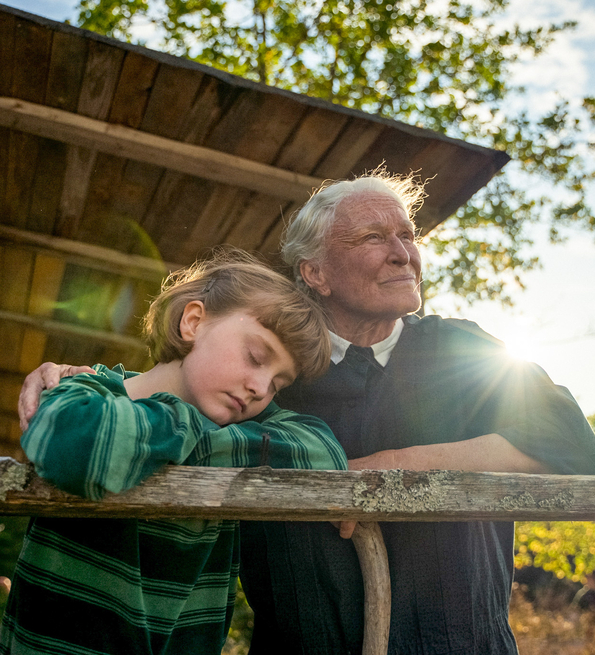 A grandmother and her granddaughter lean against a ledge on a veranda in the summer sun.