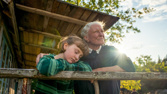 A grandmother and her granddaughter lean against a ledge on a veranda in the summer sun.