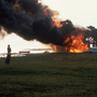 A person stands on a grassy shore watching a building engulfed in flames and thick black smoke near a parked white car.