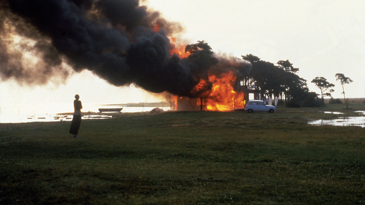A person stands on a grassy shore watching a building engulfed in flames and thick black smoke near a parked white car.