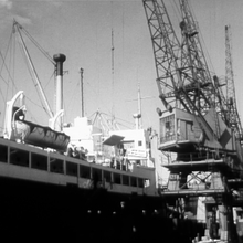 Black and white photo of a docked cargo ship with lifeboats and large cranes loading or unloading containers.