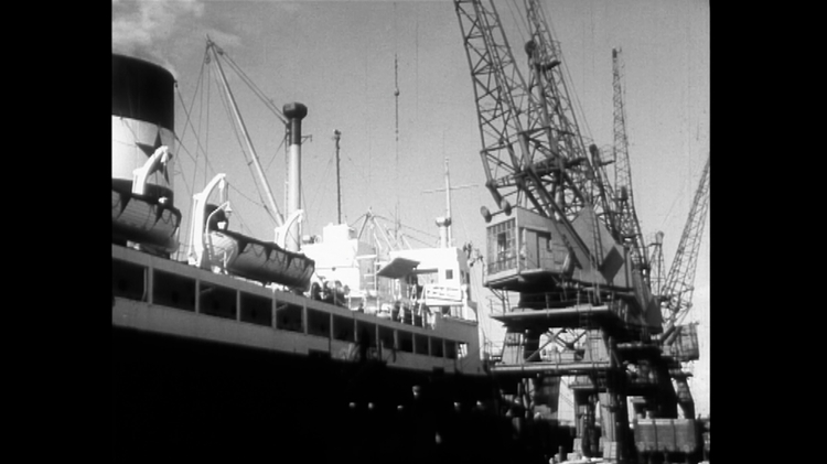 Black and white photo of a docked cargo ship with lifeboats and large cranes loading or unloading containers.