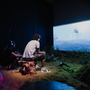 People sitting on tree stumps watching a large screen in a dimly lit room at the Undershed Gallery with grass and branches on the floor.