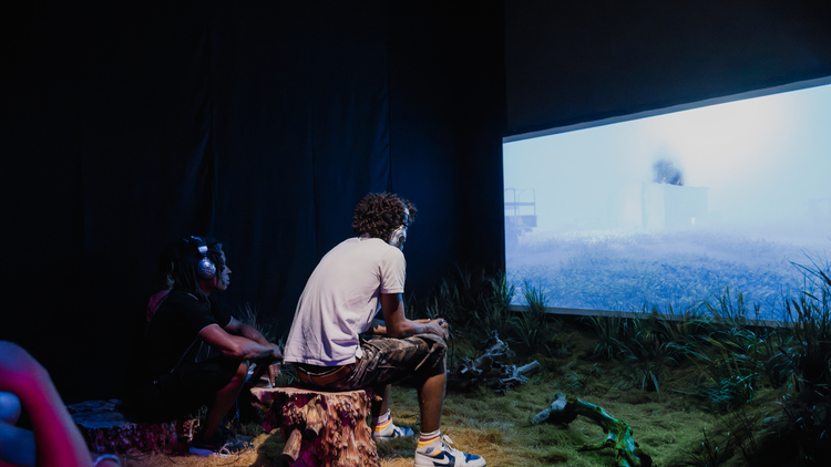 People sitting on tree stumps watching a large screen in a dimly lit room at the Undershed Gallery with grass and branches on the floor.