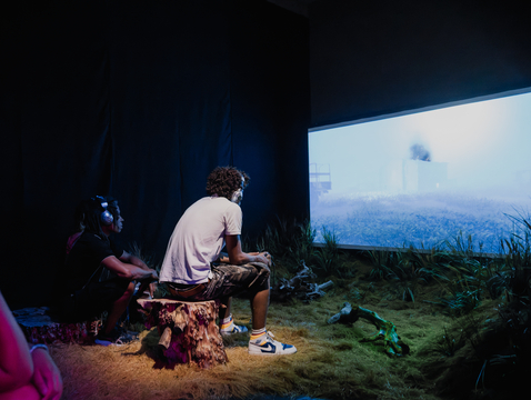 People sitting on tree stumps watching a large screen in a dimly lit room at the Undershed Gallery with grass and branches on the floor.