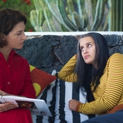 Two women sit on a striped couch outdoors; one in red reads a book while the other in yellow stripes listens attentively.