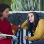 Two women sit on a striped couch outdoors; one in red reads a book while the other in yellow stripes listens attentively.