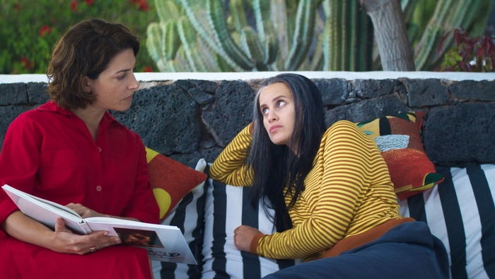 Two women sit on a striped couch outdoors; one in red reads a book while the other in yellow stripes listens attentively.