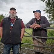 Two older men in casual outdoor clothes stand by a metal gate in a grassy rural area under a cloudy sky.