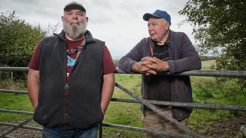 Two older men in casual outdoor clothes stand by a metal gate in a grassy rural area under a cloudy sky.