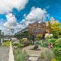 Lush urban garden with diverse plants, people sitting, historic building, river with boats, and birds flying under a bright blue sky with clouds.