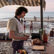 Two people facing each other under a striped canopy on a beach, with groups of people and the ocean in the background at sunset.