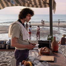 Two people facing each other under a striped canopy on a beach, with groups of people and the ocean in the background at sunset.
