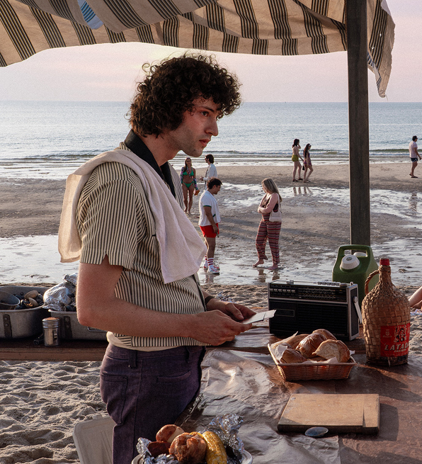 Two people facing each other under a striped canopy on a beach, with groups of people and the ocean in the background at sunset.