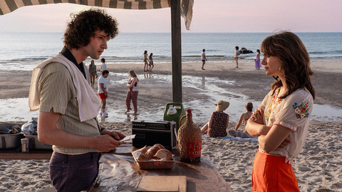 Two people facing each other under a striped canopy on a beach, with groups of people and the ocean in the background at sunset.