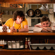 Two men lean on a wooden counter in a cozy kitchen with condiment jars, a lamp, and kitchen utensils hanging above.
