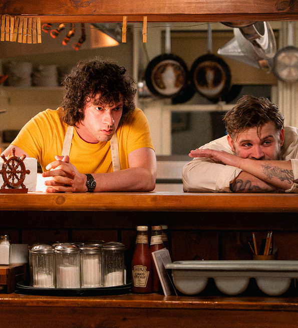 Two men lean on a wooden counter in a cozy kitchen with condiment jars, a lamp, and kitchen utensils hanging above.