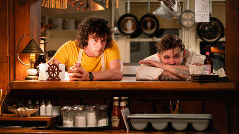 Two men lean on a wooden counter in a cozy kitchen with condiment jars, a lamp, and kitchen utensils hanging above.