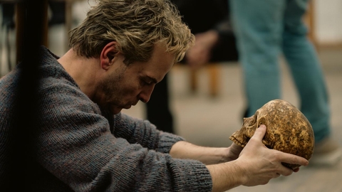 Man in a grey sweater sitting and intently looking at a weathered human skull held with both hands.