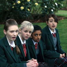 Four teenage girls in green school blazers and red-striped ties sitting on grass, looking slightly surprised or concerned.