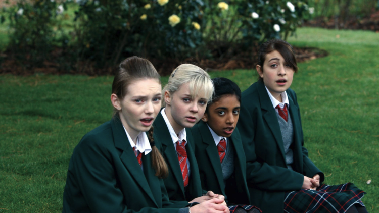 Four teenage girls in green school blazers and red-striped ties sitting on grass, looking slightly surprised or concerned.
