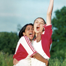 Two young women in sports jerseys celebrating joyfully outdoors with one raising a fist in the air.