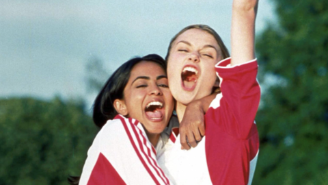 Two young women in sports jerseys celebrating joyfully outdoors with one raising a fist in the air.