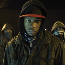 Group of five hooded individuals wearing masks and jackets, standing outdoors at night with serious expressions.
