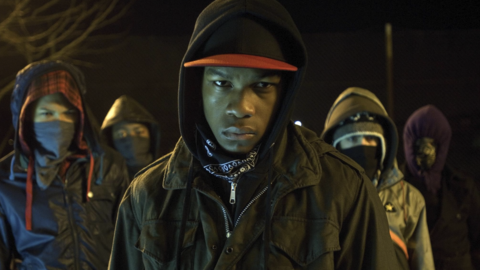 Group of five hooded individuals wearing masks and jackets, standing outdoors at night with serious expressions.