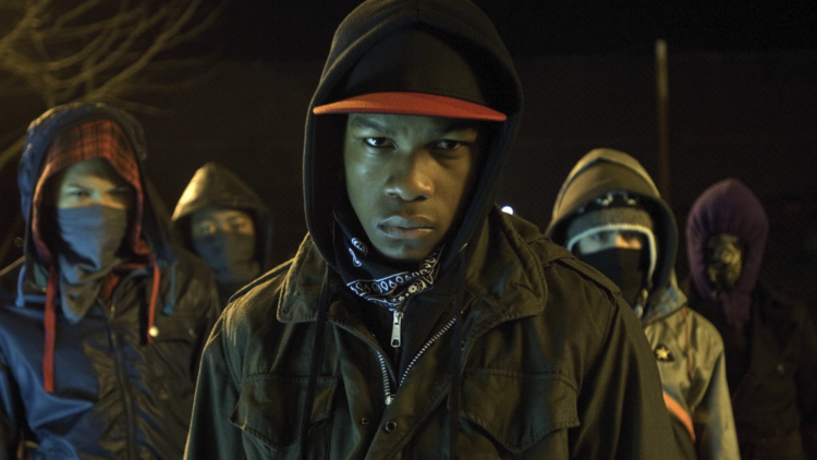 Group of five hooded individuals wearing masks and jackets, standing outdoors at night with serious expressions.