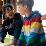 Smiling woman and boy collaborate at a table with a laptop and craft supplies in a bright classroom, creating a warm, engaging scene.