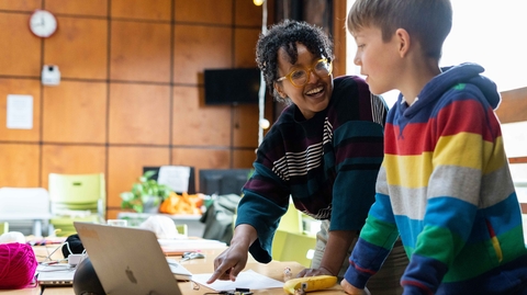 Smiling woman and boy collaborate at a table with a laptop and craft supplies in a bright classroom, creating a warm, engaging scene.