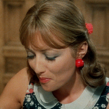 Woman in floral dress with red earrings and hair accessory eating with a spoon, seated in a vintage wooden chair.