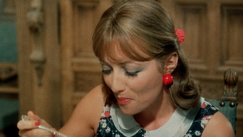Woman in floral dress with red earrings and hair accessory eating with a spoon, seated in a vintage wooden chair.