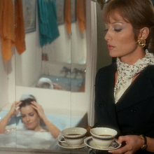 Woman in a black suit holding two cups of coffee in a bathroom while another woman relaxes in a bubble bath in the background.