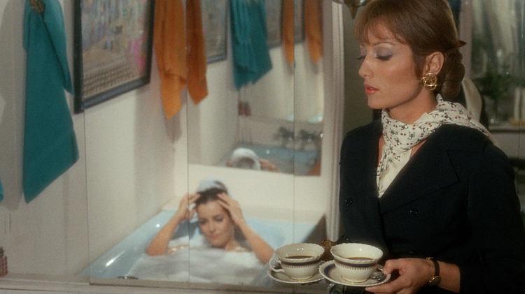 Woman in a black suit holding two cups of coffee in a bathroom while another woman relaxes in a bubble bath in the background.
