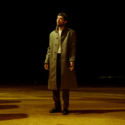 Man in a long grey coat standing alone on a dimly lit empty road at night with a dark sky background.