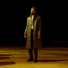 Man in a long grey coat standing alone on a dimly lit empty road at night with a dark sky background.