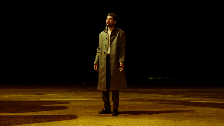 Man in a long grey coat standing alone on a dimly lit empty road at night with a dark sky background.